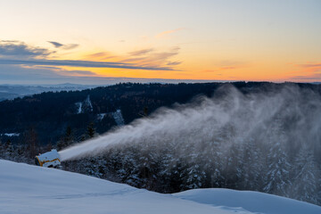 Snow cannon operates on a mountain slope, creating artificial snow for skiing. The sun sets in the background, adding color to the winter landscape. Trees are covered in snow.