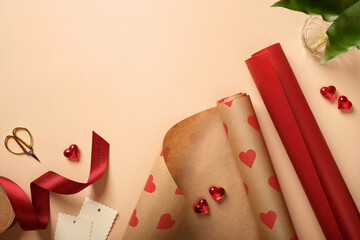 Gift wrapping supplies with red paper rolls, ribbon, scissors, tag and heart decorations on beige background. Copyspace.