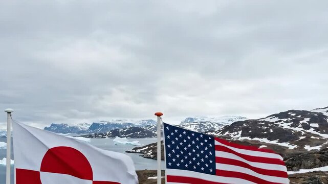 The image shows the flags of Greenland and the United States waving in a cold, coastal landscape.  The background features a body of water with icebergs, snowy mountains, and rocky terrain.