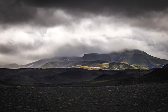 View of stark, dark volcanic ground stretches toward misty mountains, where sunlight kisses the green slopes under a brooding sky, Landmannalaugar, The Central Highland, Iceland.
