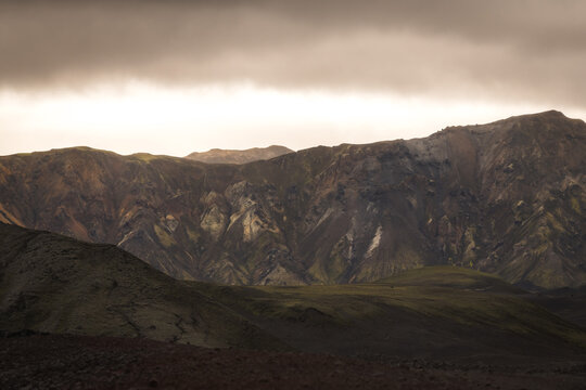 View of rugged peaks rise under a moody sky, their slopes painted with earthy hues, creating a raw, untouched landscape, Landmannalaugar, The Central Highland, Iceland.
