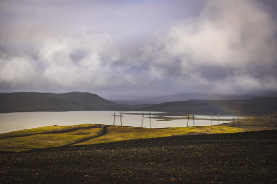 View of golden light painting the grassy banks of a serene lake beneath a brooding sky, power lines marching across the horizon, Landmannalaugar, The Central Highland, Iceland.
