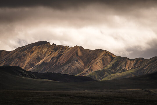 View of rugged, multi-colored mountains rise under a brooding sky, casting dramatic shadows across the Icelandic highlands, Landmannalaugar, The Central Highland, Iceland.