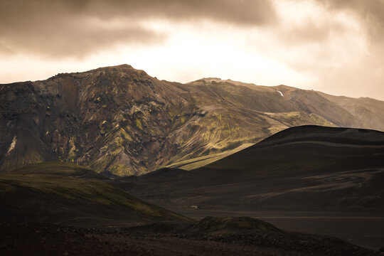 View of jagged, dark mountains rise against a stormy sky, their slopes marked with streaks of green, contrasting with the shadowed foreground, Landmannalaugar, The Central Highland, Iceland.