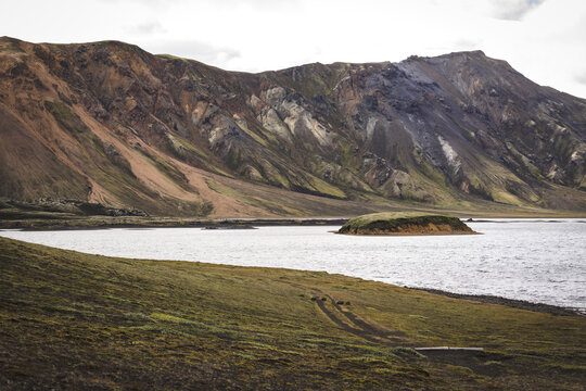 View of rugged, multicolored mountains rise majestically above a serene lake reflecting the cloudy sky, with a small island in the distance, Landmannalaugar, The Central Highland, Iceland.