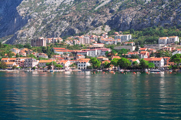 Fototapeta premium Scenic coastline of village of Dobrota in Bay of Kotor, Montenegro, at base of steep mountains. Traditional red roofed buildings line water edge, contrasting with blue sea and rugged natural landscape