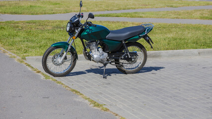Obraz premium A green motorcycle is parked on a sidewalk beside a grassy area during the daytime. The bike sits on a flat surface with some sunlight