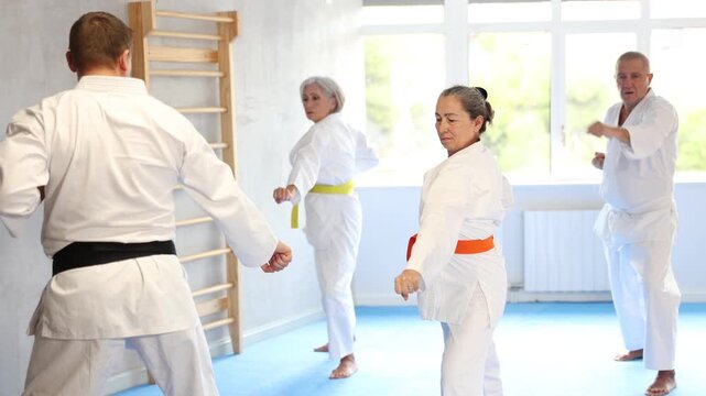Diligent old man attendee of karate classes practicing kata standing in row with others in sports hall