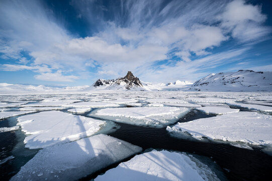 A vast, icy landscape under a dramatic sky. Jagged mountains rise in the distance, their peaks dusted with snow, while the foreground is a mosaic of dark water and broken ice floes.