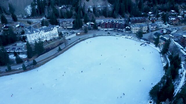 Aerial drone footage of a frozen lake in Champex, Switzerland, during winter. People ice skating surrounded by snowy alpine scenery. Shot in 4K using a DJI Mini 2.