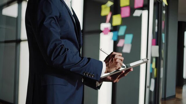 businessman using digital tablet with stylus, standing in modern office, blurred glass board with colorful sticky notes in background