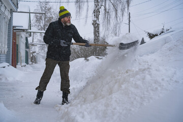 A man in a dark jacket shovels snow near a house, a village street, winter, snowdrifts