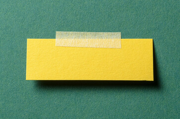 Piece of blank yellow paper note with adhesive sticky tape and shadow on green background