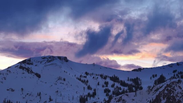 Dramatic sunset clouds swirl above snow covered mountain slopes and dark pine trees. Winter alpine landscape with glowing evening sky, cold atmosphere and peaceful mood in California, USA
