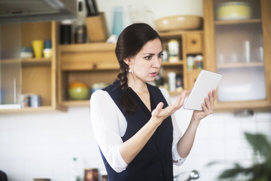 Side view of a serious Woman standing in her kitchen video conferencing on a digital tablet