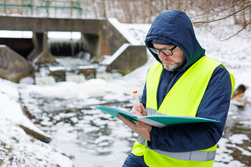 Male ecologist collected a water sample while standing near a sluice gate in a wintry forest. He recorded information about the meltwater level in the lake.