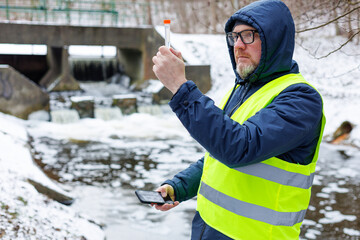 Male ecologist holds a test tube containing a water sample. He collected a meltwater sample for analysis near a sluice gate in a snow covered forest during winter.