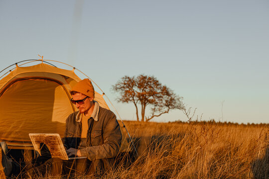 Male digital nomad using computer and working online while sitting near camping tent at campsite in nature  