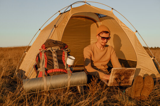 Male traveler browsing laptop during remote work while sitting near camping tent at campsite in nature at sunset  