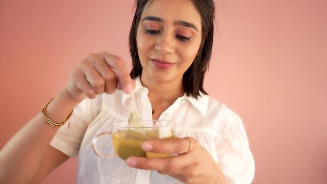 Close-up head shot of happy relaxed pretty young Asian woman girl hold cup dipping teabag, preparing herbal green tea with calm face expression, isolated over pink background.