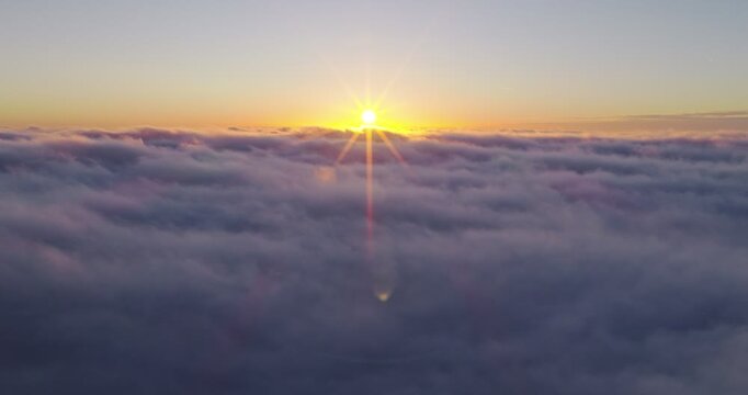 Aerial flight above glowing clouds at sunset with golden sunlight breaking through. Cinematic skyscape with soft fog, warm colors and dramatic atmosphere high above the horizon.