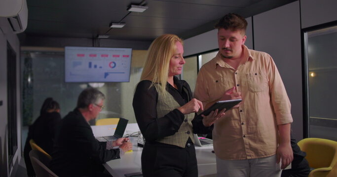 Young man and blonde businesswoman collaborating over digital tablet in office, team working in background with presentation data on monitor - Powered by Adobe
