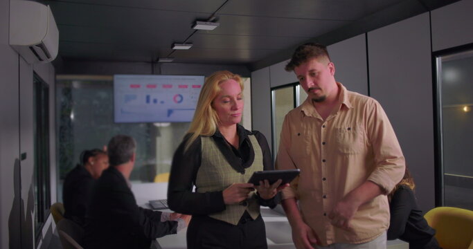 Young man and blonde businesswoman collaborating over digital tablet in office, team working in background with presentation data on monitor - Powered by Adobe