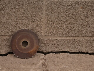 Old rusty circular saw blade leaning against concrete base near gray wall, industrial look.