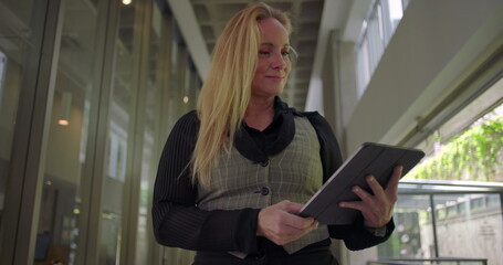 Woman using tablet with gentle smile in modern hallway, interacting with digital screen while appearing satisfied and engaged in work or communication