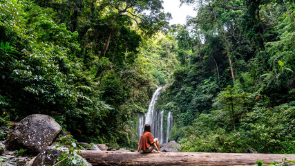 Young woman sitting on a fallen log in front of a waterfall in the jungle