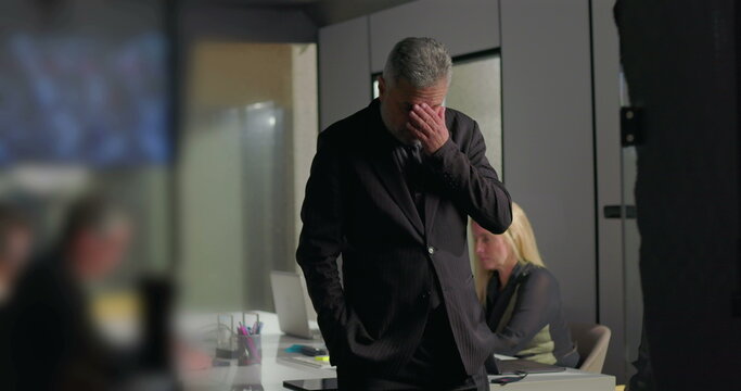 Worried businessman covering face with hand while walking across office conference room during tense boardroom meeting with coworkers in background