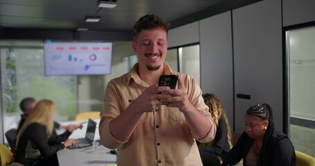 Young businessman smiling at smartphone during meeting, happy reaction while colleagues work at laptops in modern conference room with data screen