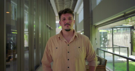Young man standing in modern office hallway with neutral expression, hands in pockets, confident and relaxed posture in corporate environment