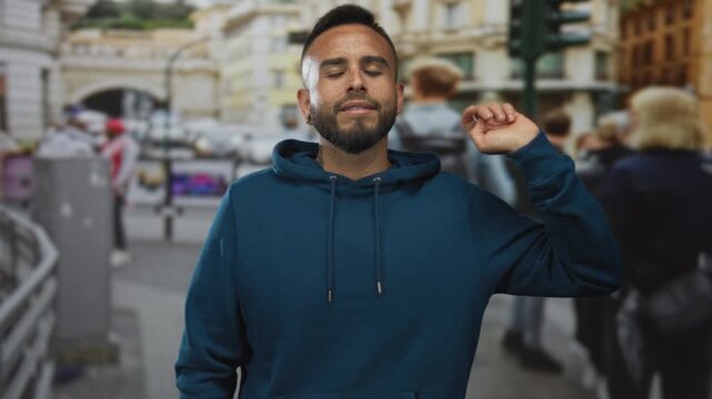 Man showing three fingers with raised hand, beard visible and blue hoodie, standing on busy city street by traffic lights and buildings; confidence casual.
