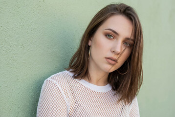 Young woman with striking green eyes and hoop earrings poses against a textured green wall