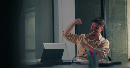 Young man throws arms up in joyful celebration after successful moment at laptop during solo office work session