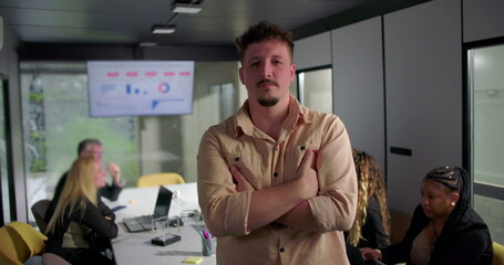 Young man with arms crossed looking determined in office setting, colleagues working in background during meeting with data visualization on screen