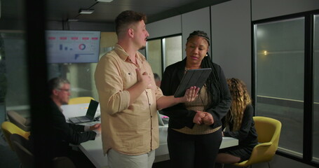 Young man and African American businesswoman discussing digital content on tablet in office, standing near team working at conference table with data screen