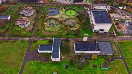 Aerial view shows an abandoned industrial area with multiple buildings, circular tanks, and overgrown vegetation. The scene is captured on a cloudy day, revealing the neglect over time.