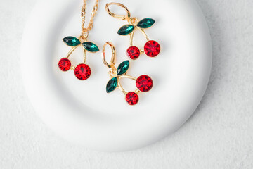 Elegant cherry earrings and necklace set with red gemstones and green leaves placed on white round...
