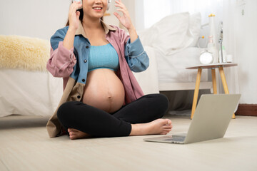 A pregnant woman sits cross-legged on the floor with headphones, engaging with a laptop in a cozy bedroom. The scene highlights modern prenatal lifestyle, digital learning, and maternal wellbeing