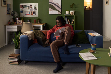 Young Black man sitting on sofa multitasking with laptop and paperwork, focusing on screen while working from home in living room, smartphone and documents nearby