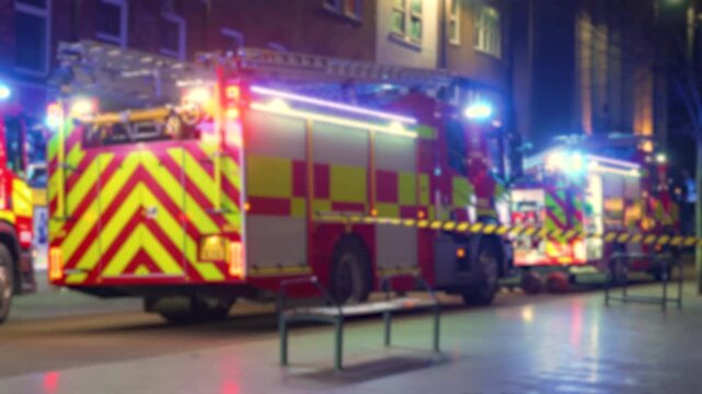 Firefighting vehicles blocking night street during active emergency with flashing lights. Several rescue engines standing stationary near buildings amid urgent incident. Multiple emergency trucks