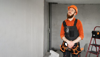 Male bearded builder in hard hat and blue overalls with working tools on belt smiling and looking...