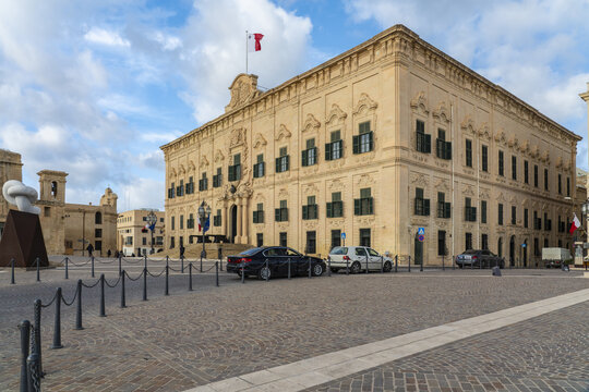 View of imposing Il-Ber&Auml;&iexcl;a ta' Kastilja building rise above the textured square while cars sit parked, and a flag flies high against a blue, lightly clouded sky, Valletta, Malta.