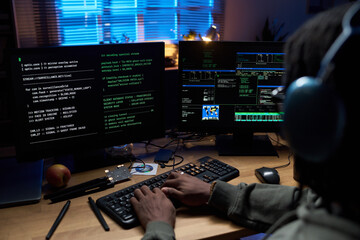 Black young man wearing headphones working on computer with multiple monitors displaying programming code and cybersecurity data in dark office at night