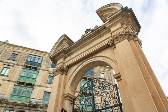 View of a decorative stone archway framing a glimpse of buildings with green balconies against a cloudy sky, creating a captivating architectural tableau, Valletta, Malta.