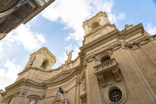 View of the majestic baroque architecture of St. Catherine of Italy Church rises towards a sky streaked with wispy clouds, bathed in warm sunlight, Valletta, Malta.