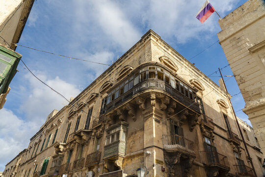 View of weathered, pale yellow stone buildings rise against a bright blue sky with a Russian flag fluttering above, creating a striking contrast of textures and colors, Valletta, Malta.