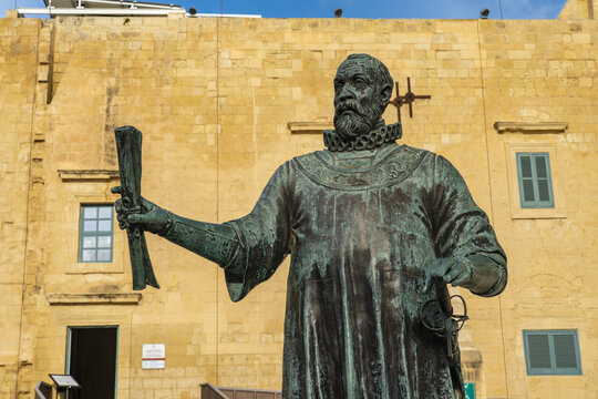 View of bronze statue of a man holding a scroll stands proudly against a weathered yellow stone building with small windows, Valletta, Malta.